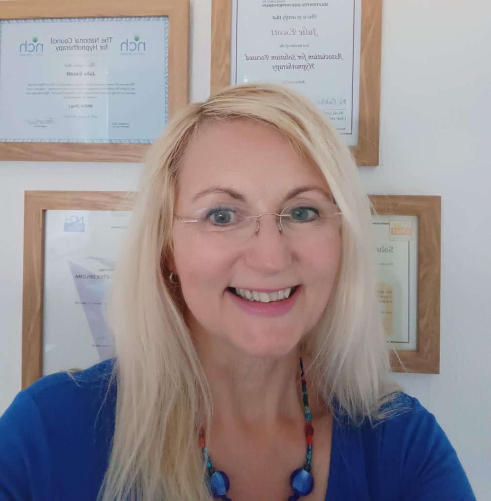 portrait of julie escott, blonde woman with glasses in front of certificates wall